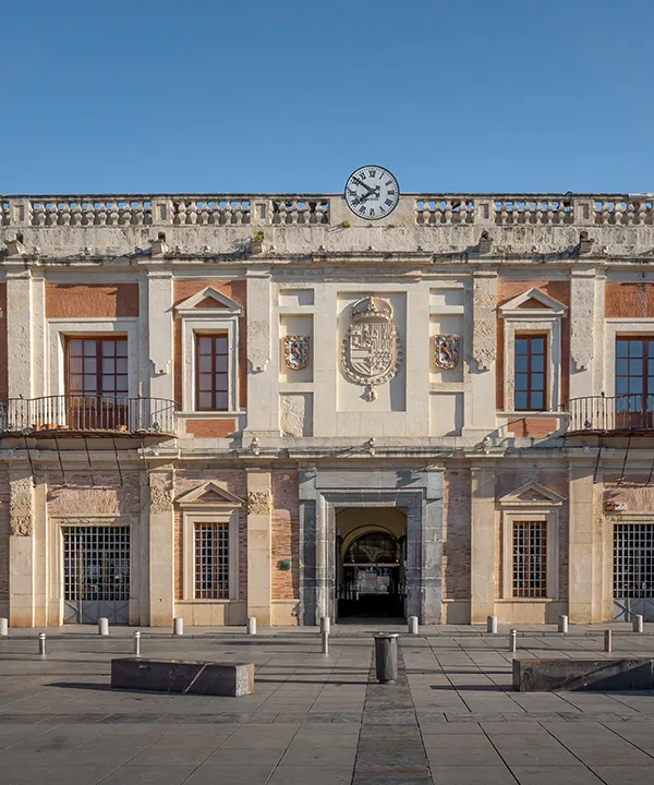 Edificio hist&oacute;rico de fachada sim&eacute;trica con reloj y escudo bajo un cielo azul.