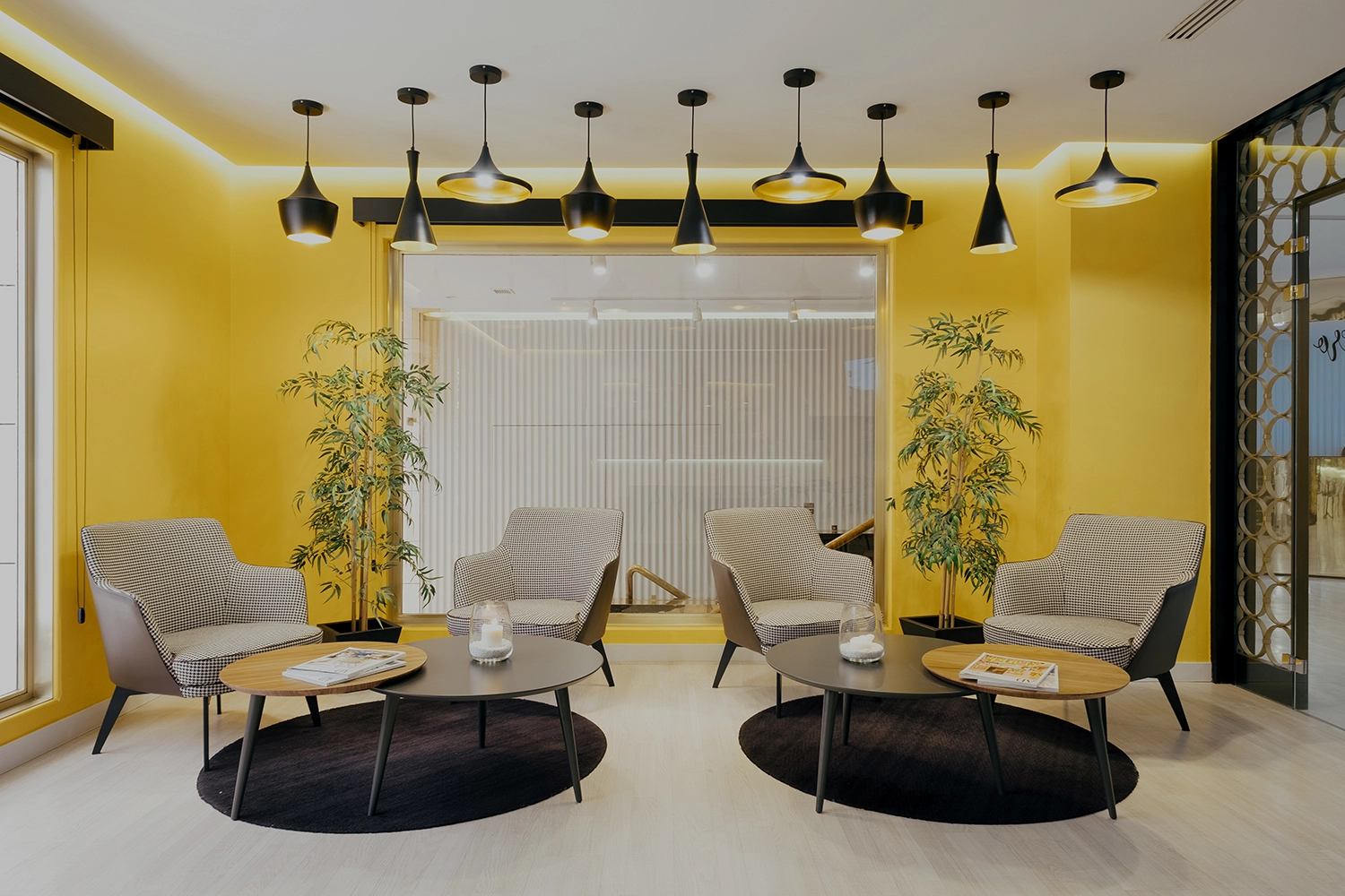 Sala moderna con sillones, mesas redondas y plantas en paredes amarillas.