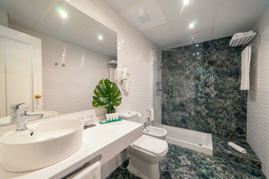 Baño moderno con lavabo, bidé y ducha, decorado con planta y mármol oscuro.