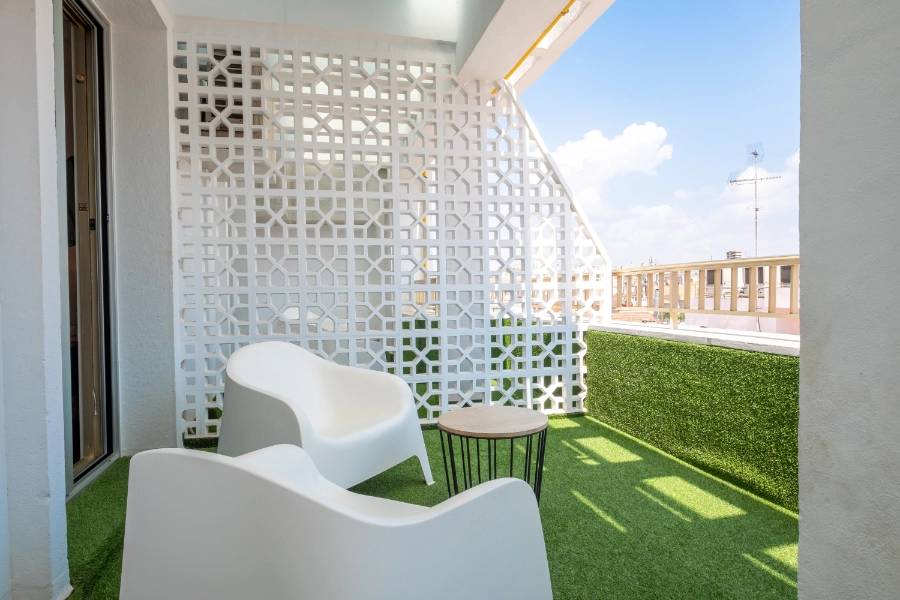 Balcón con sillas blancas, mesa y celosía decorativa, vista al cielo despejado.