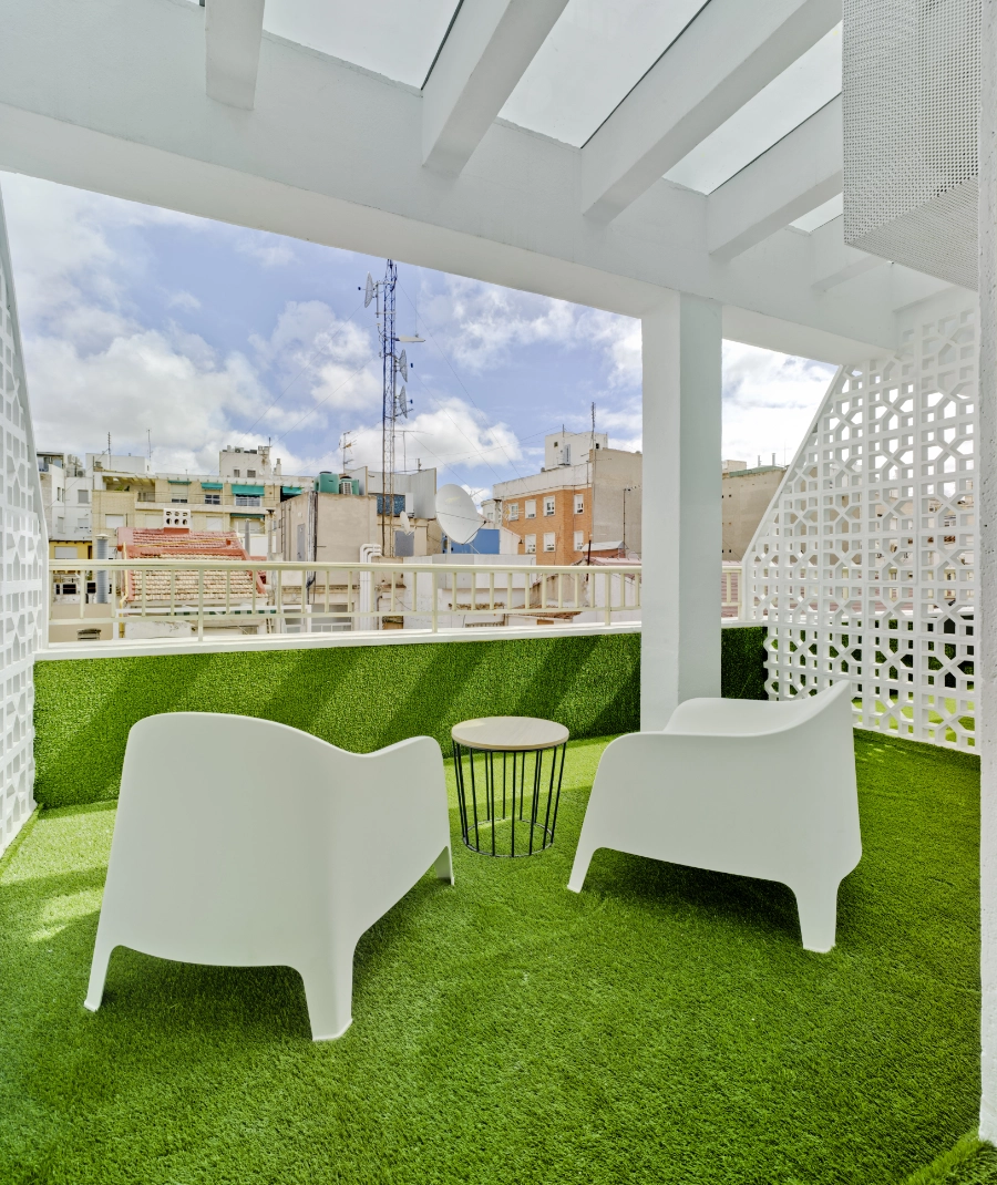 Terraza con sillas blancas y césped artificial, vista de edificios al fondo.