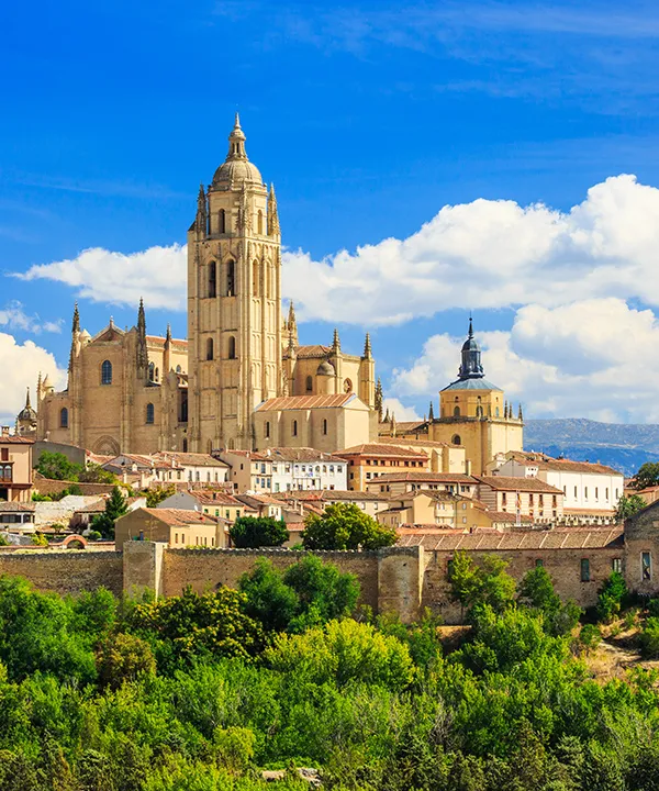 Catedral de Segovia bajo un cielo azul con nubes, rodeada de edificios y vegetaci&oacute;n.
