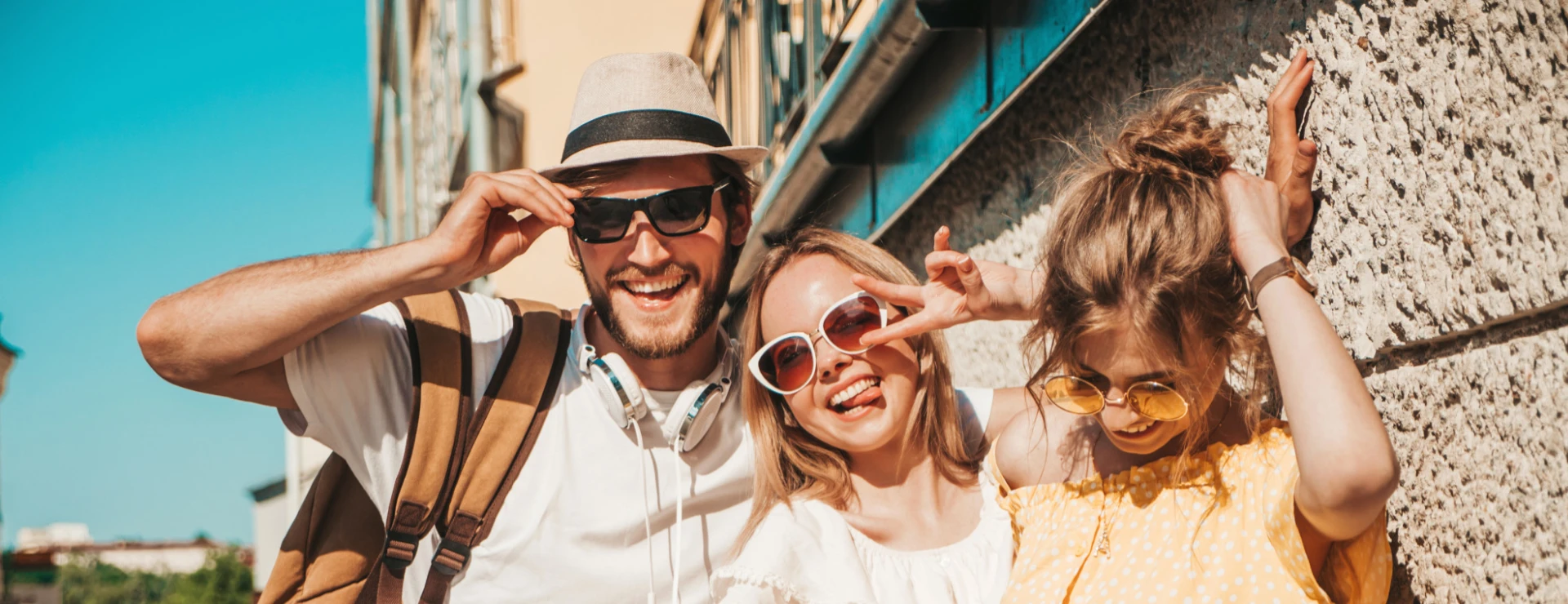 Tres personas sonriendo con gafas de sol y ropa veraniega junto a una pared.