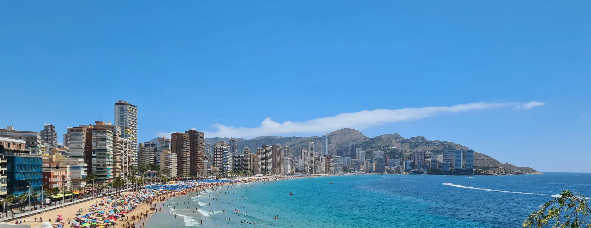 Playa llena de gente, con rascacielos y monta&ntilde;as al fondo bajo un cielo azul.
