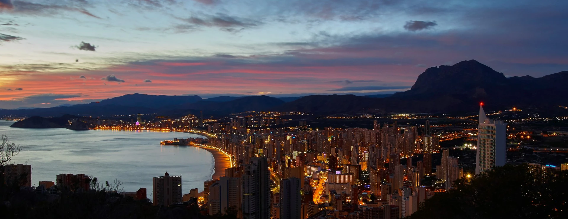 Ciudad costera iluminada al anochecer con monta&ntilde;as al fondo.