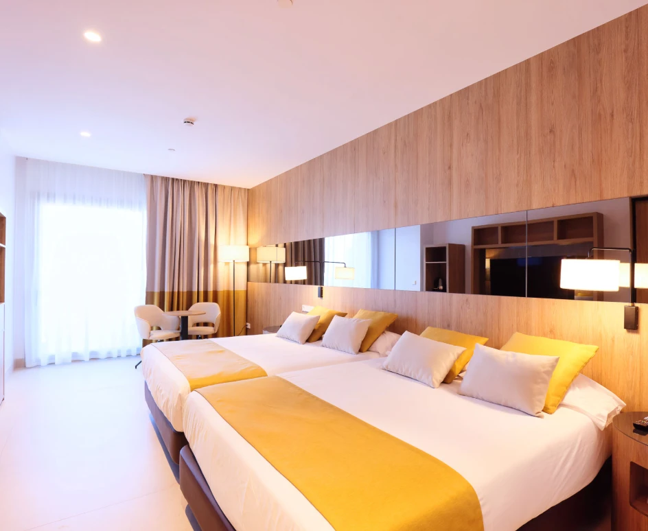 Habitaci&oacute;n moderna con dos camas, tonos c&aacute;lidos y ventana amplia con cortinas.