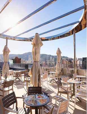 Terraza soleada con mesas, sillas y sombrillas cerradas, vista urbana de fondo.