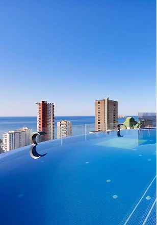 Piscina infinita con vista al mar y edificios al fondo bajo cielo azul.