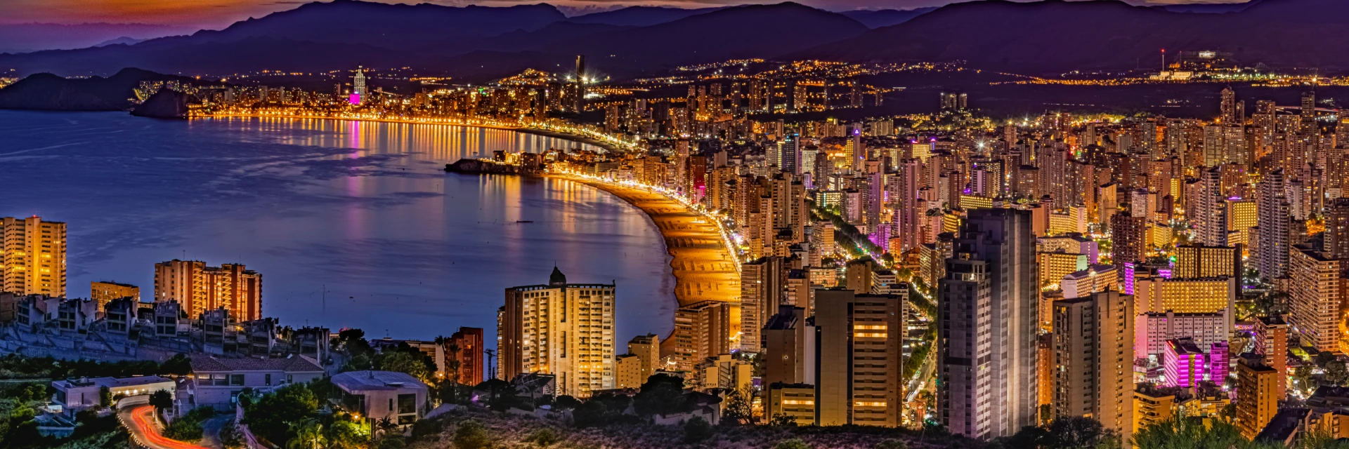 Ciudad costera iluminada de noche, con un skyline moderno y edificios altos.
