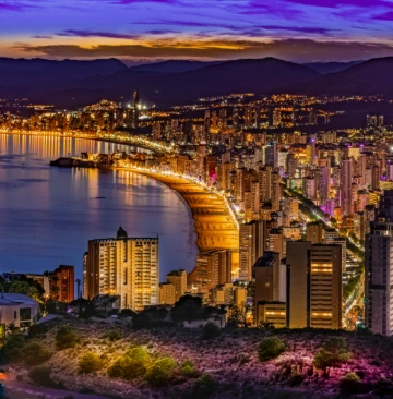 Ciudad costera iluminada al anochecer con un cielo p&uacute;rpura y monta&ntilde;as al fondo.