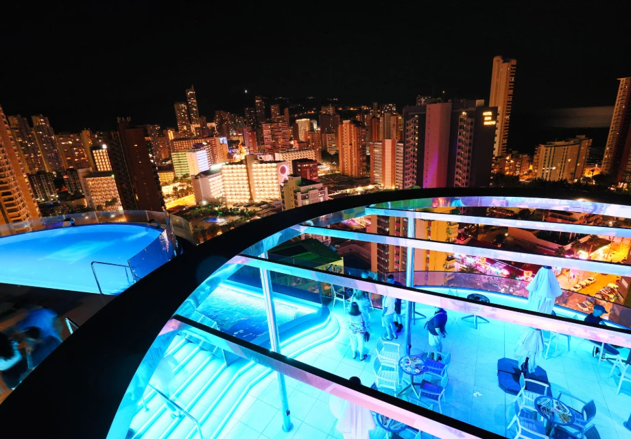 Vista nocturna de una ciudad con edificios y piscina iluminada en una azotea.