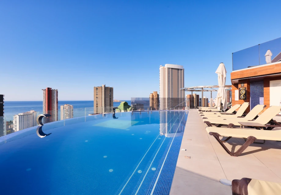 Piscina infinita en azotea con vista al mar y rascacielos en el fondo.
