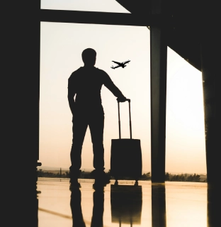 Hombre con maleta observa un avi&oacute;n despegando en el aeropuerto al atardecer.