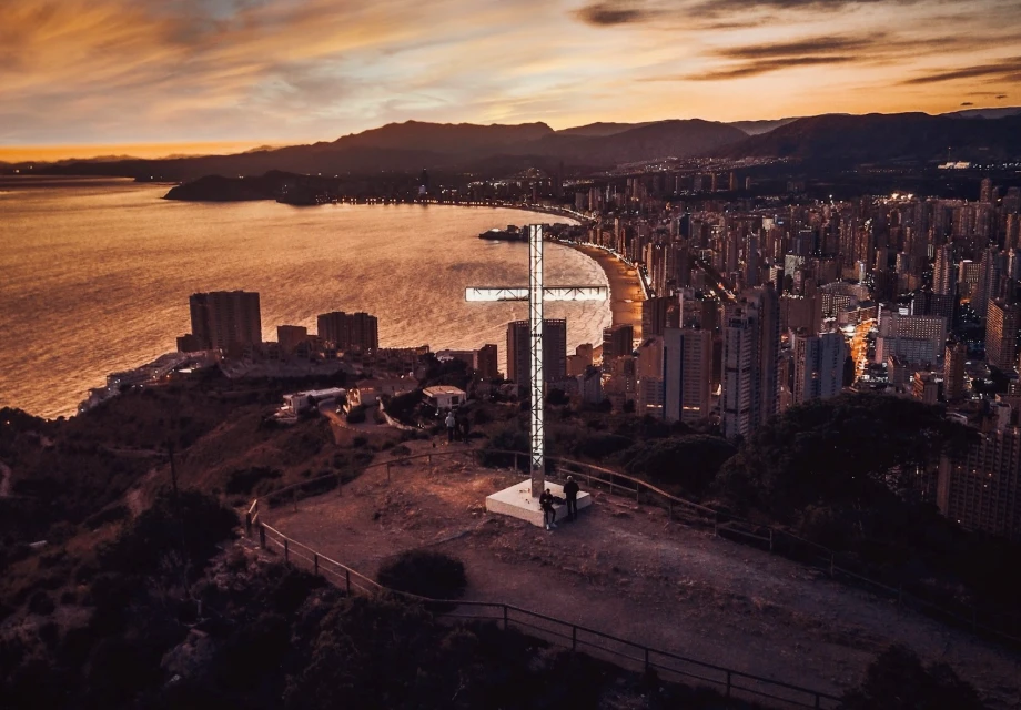 Cruz iluminada sobre la ciudad al anochecer con la costa al fondo.