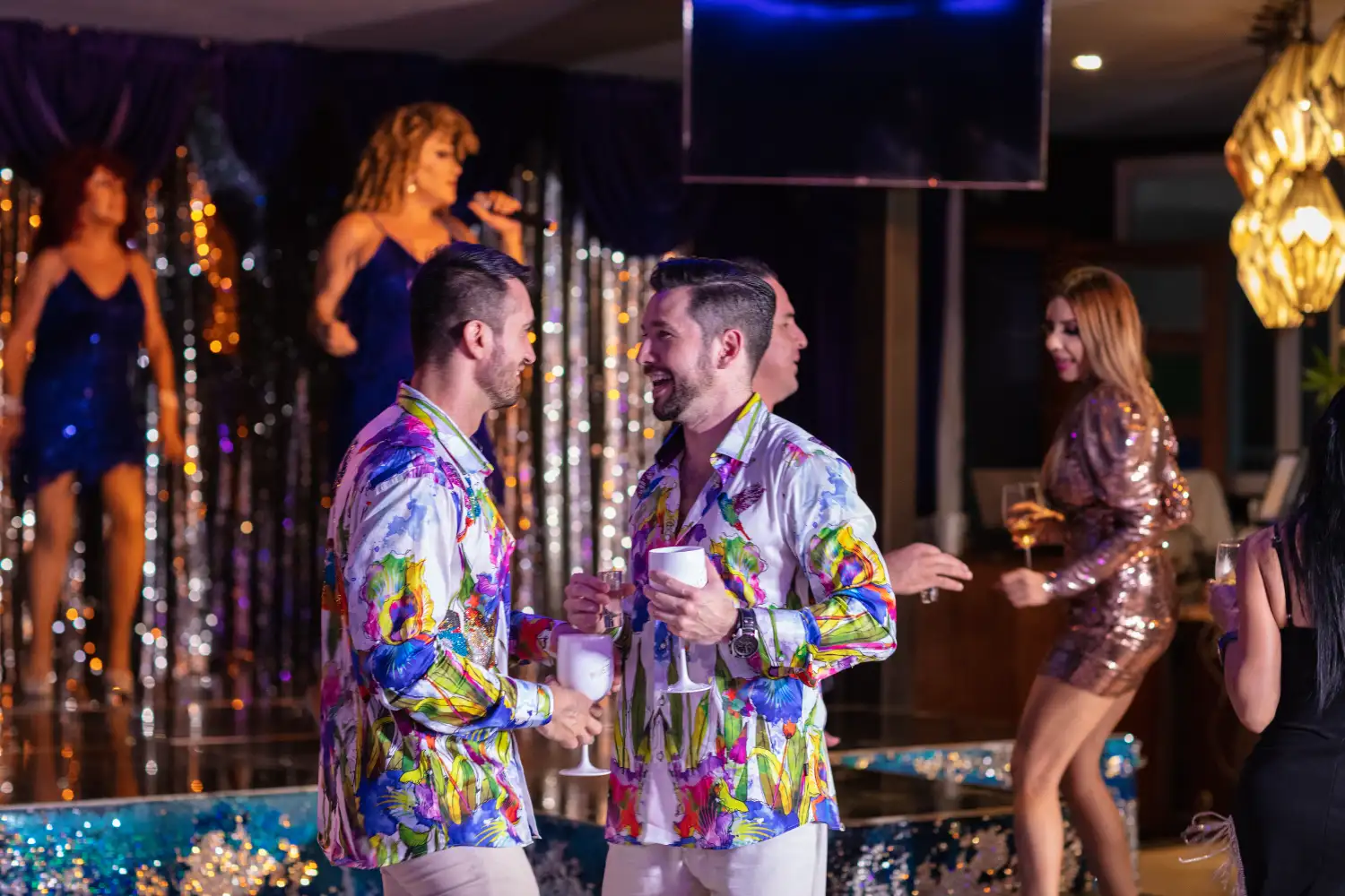 Personas bailando en una fiesta con trajes coloridos y espect&aacute;culo en el fondo.