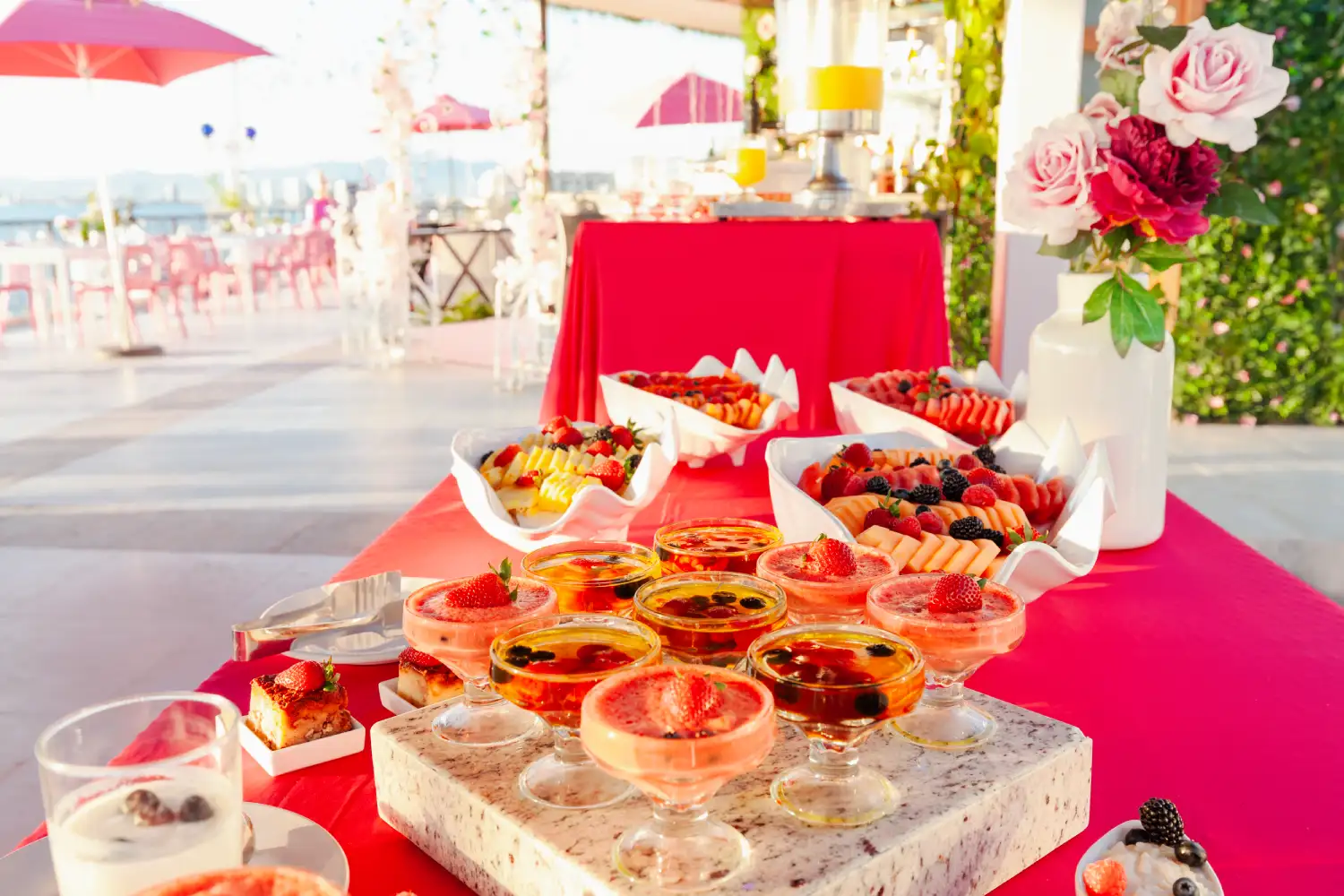 Mesa de postres coloridos con frutas y flores en un entorno al aire libre.