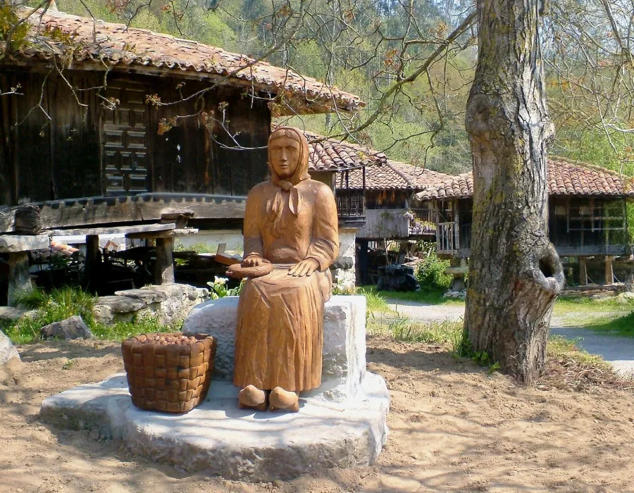 Estatua de bronce con vestimenta tradicional en parque rural, junto a casas y &aacute;rbol