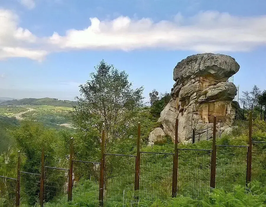 Gran roca en un paisaje verde con cielo azul y una cerca en primer plano.