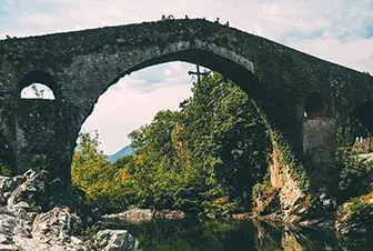 Puente de piedra antiguo rodeado de vegetaci&oacute;n y reflejado en el agua.