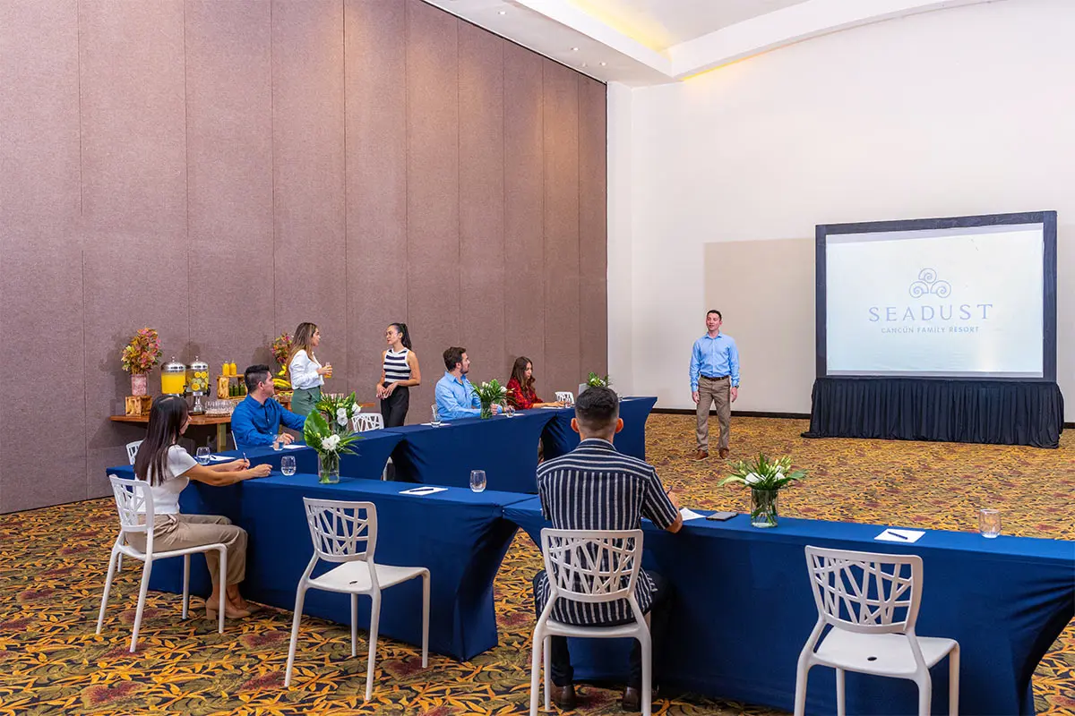 Conference room with people seated at blue tables and a presenter onstage