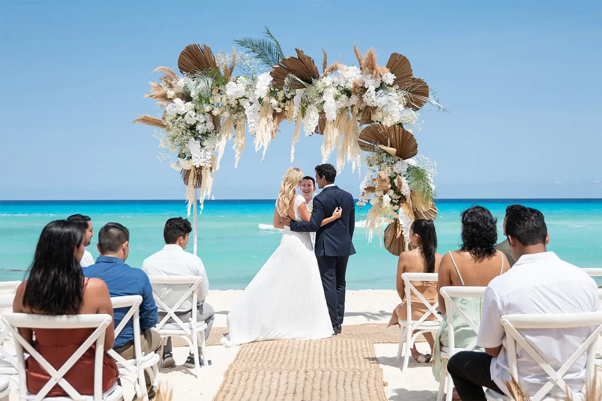Beach wedding ceremony with couple under a white floral arch by the sea