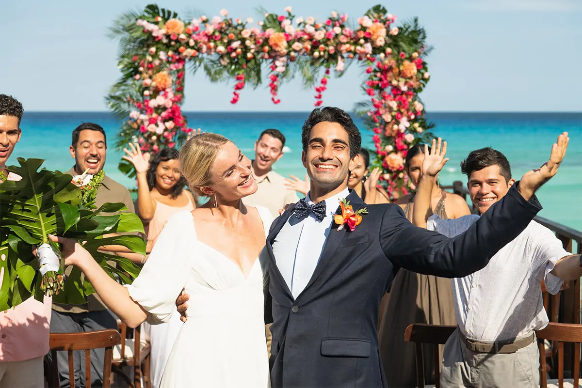 Smiling groom in tuxedo waves at beach wedding with floral arch and guests