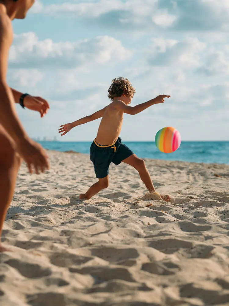 Boy running on a sunny beach, chasing a colorful ball, with ocean behind him
