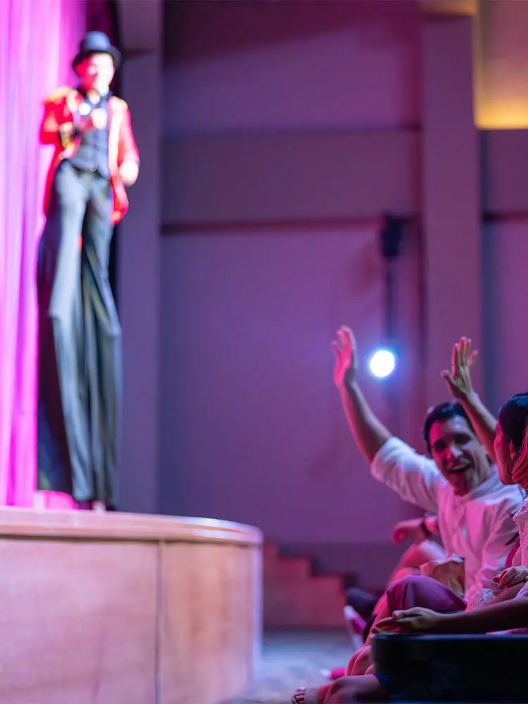 Performer on stage with raised hands as audience applauds under purple lights