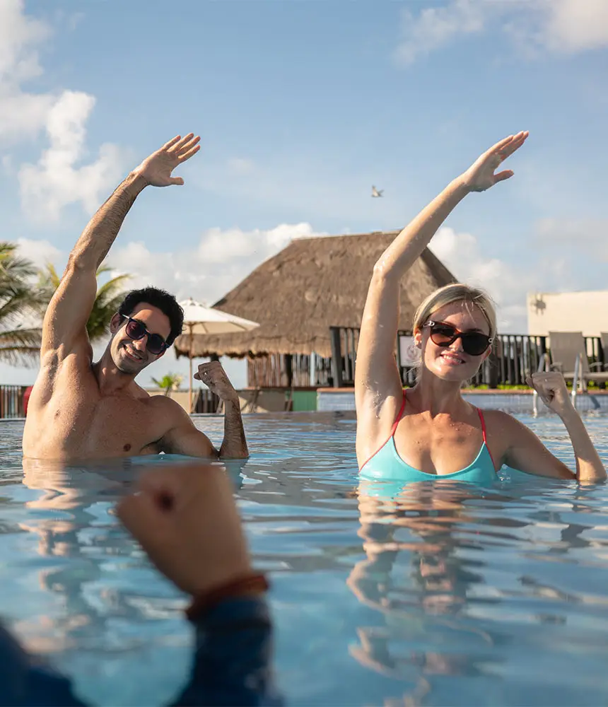 Couple in a sunny pool throwing their hands up while smiling and wearing sunglasses