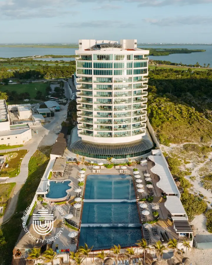 Aerial view of a beachfront hotel with pools, palms, and oceanfront dunes.