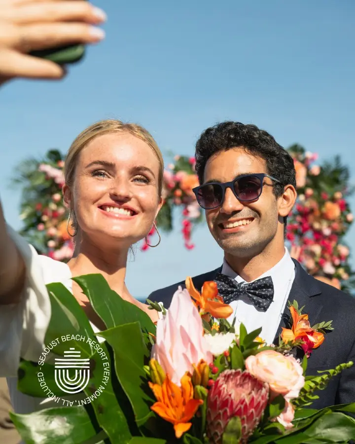 Couple in formalwear at beach event holding bouquets and smiling in sun