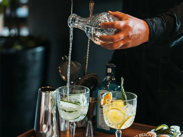 Hand pours clear liquid into glass over bar with citrus garnish and ice