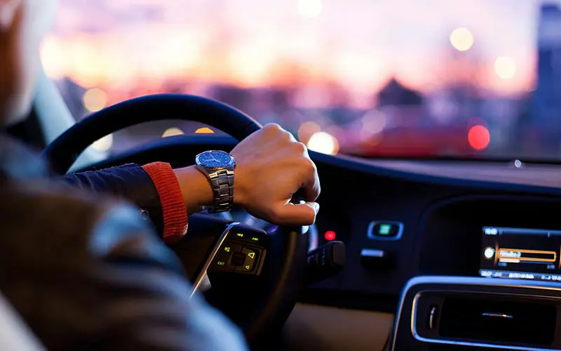 Person driving car at dusk, gripping steering wheel with dashboard lights on.