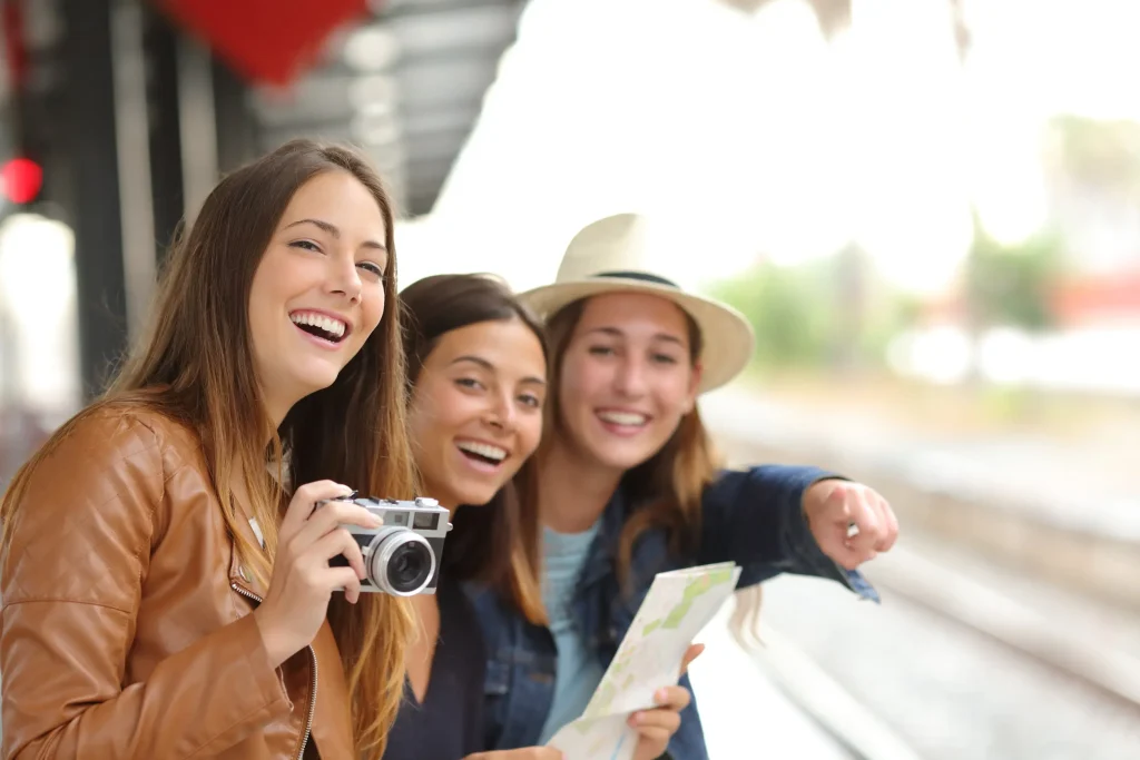 Tres mujeres sonrientes con c&aacute;mara y mapa en una estaci&oacute;n de tren.