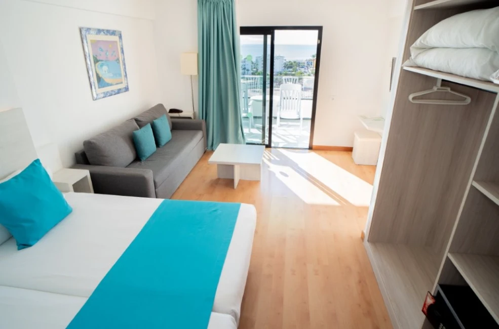 Habitaci&oacute;n de hotel luminosa con cama, sof&aacute;, cortinas azules y acceso a un balc&oacute;n con sillas.
