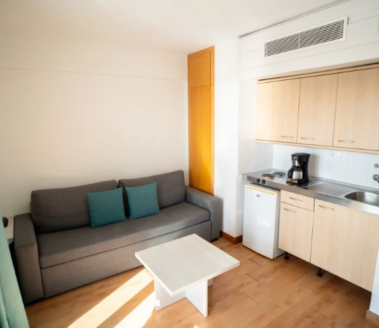 Sala con sof&aacute; gris, mesa blanca, peque&ntilde;a cocina con muebles claros, fregadero y cafetera.