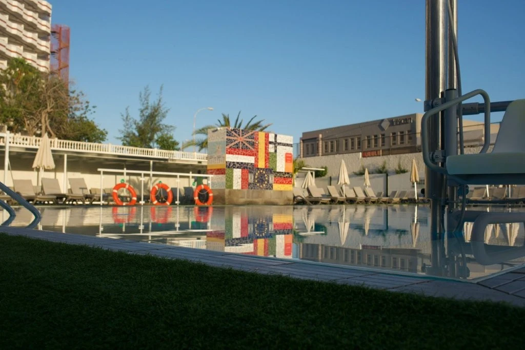 Piscina con sillas, sombrillas y un dise&ntilde;o de banderas en la pared al fondo.