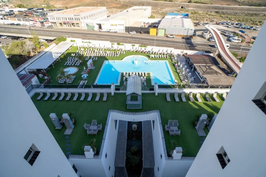 Vista a&eacute;rea de una piscina rodeada de tumbonas y c&eacute;sped, con carretera y edificios al fondo.