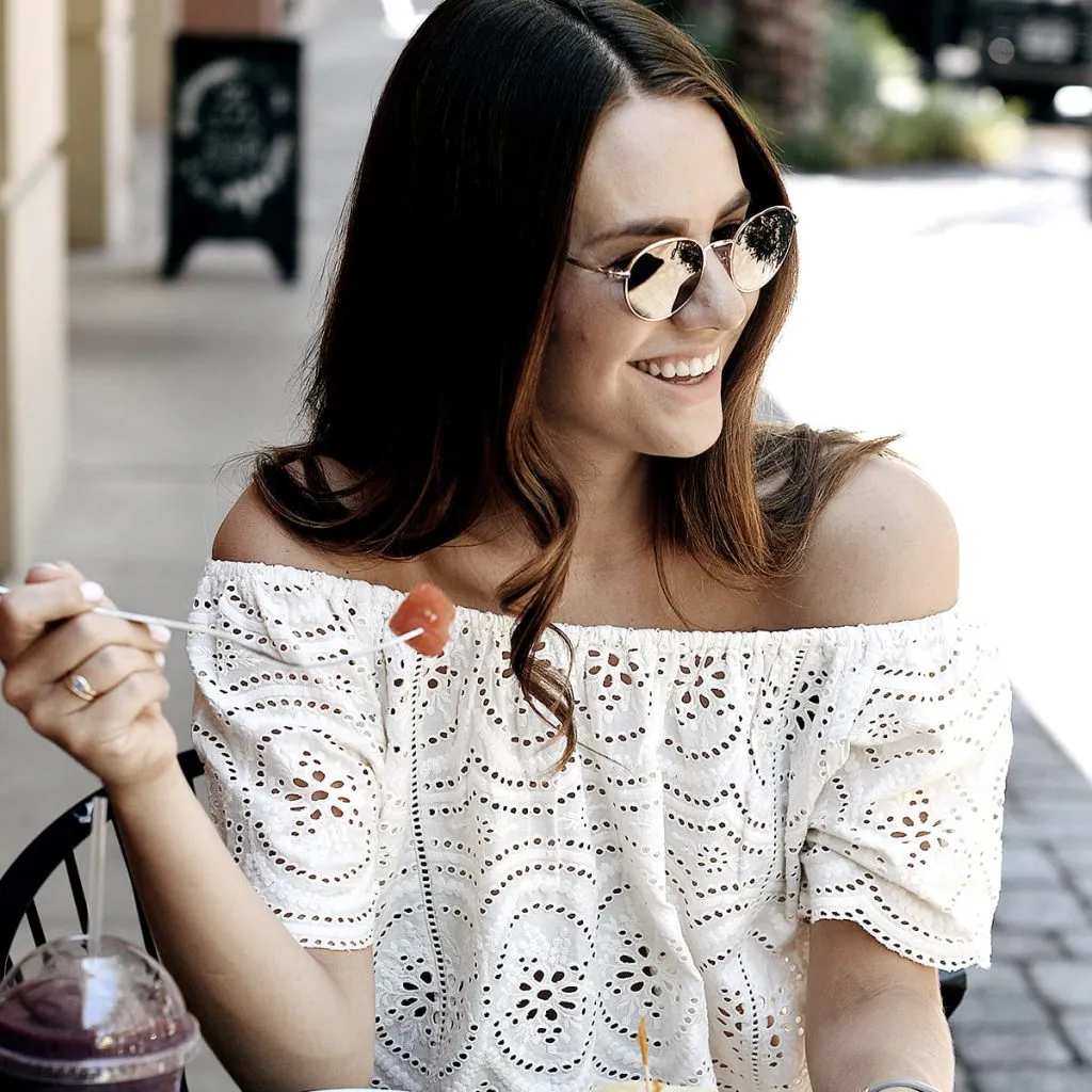 Mujer sonriente con gafas de sol y blusa blanca, tomando un bocado