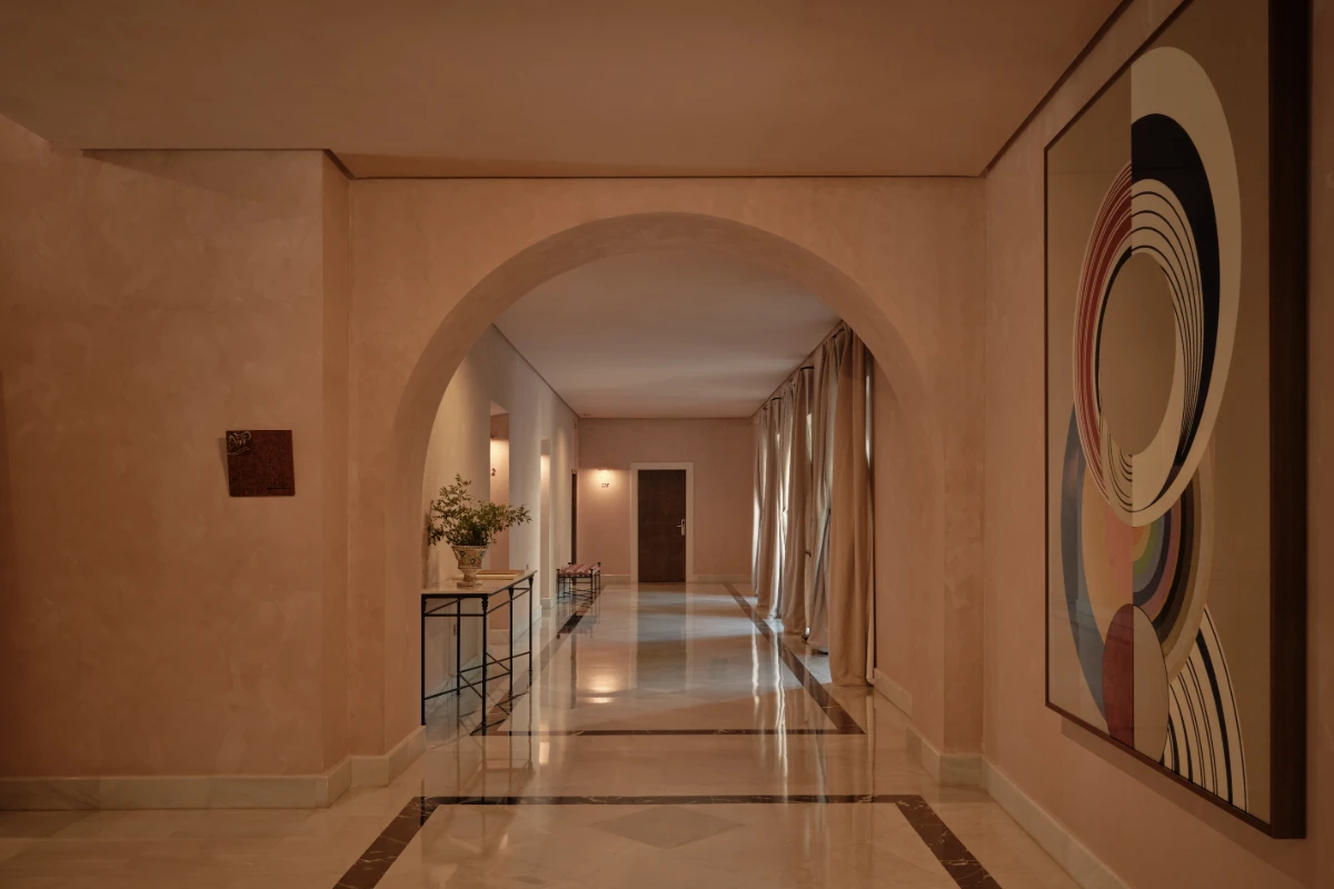 Pasillo de galer&iacute;a con arcos, cuadros y cortinas, iluminado con luz c&aacute;lida.