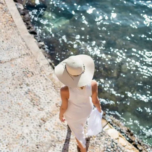 Mujer con vestido y sombrero camina junto al mar, mirando al agua