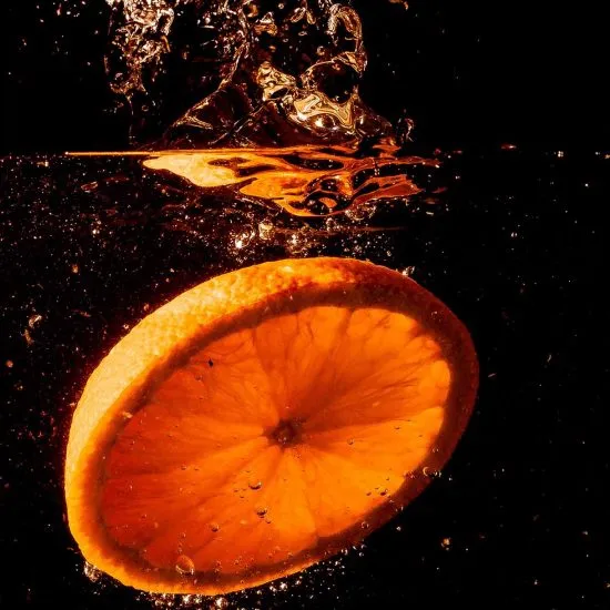 Rodaja de naranja con salpicaduras y chorros de l&iacute;quido en fondo negro