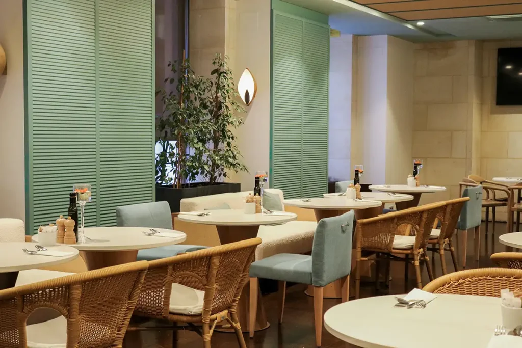 Restaurante con mesas redondas, sillas de mimbre y persianas verdes cerradas.