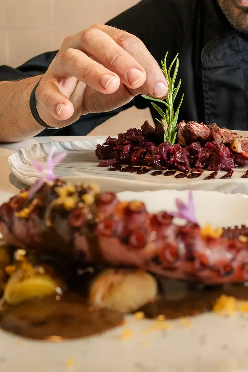 Mano colocando romero sobre carne con guarniciones en una bandeja
