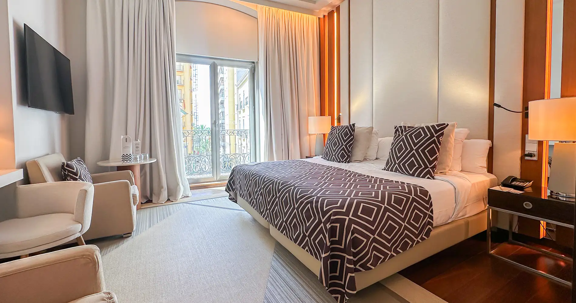 Habitaci&oacute;n de hotel con cama, sillones y cortinas junto a balc&oacute;n.