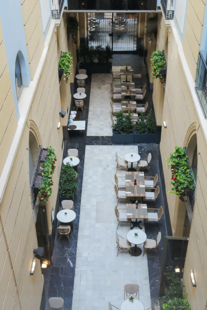 Pasillo interior de hotel con mesas, sillas y plantas decorativas.