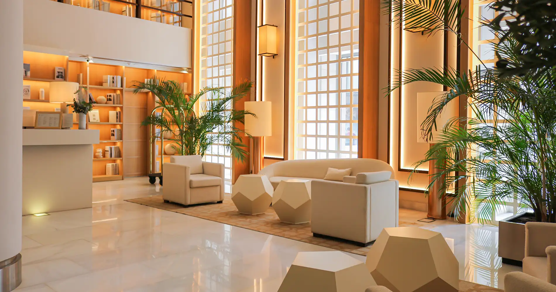 Lobby moderno con sof&aacute;s blancos, palmeras y luz c&aacute;lida junto a ventanales