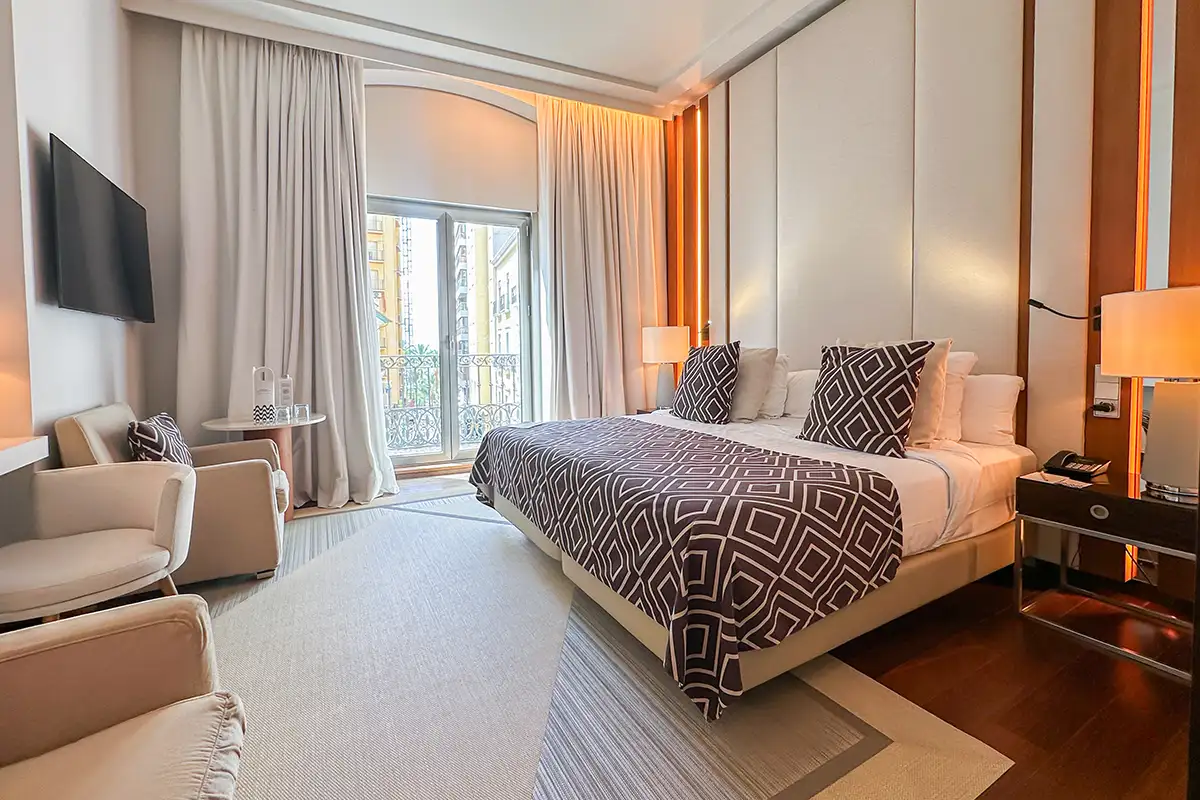 Habitaci&oacute;n de hotel moderna con cama doble, sillones y cortinas al balc&oacute;n