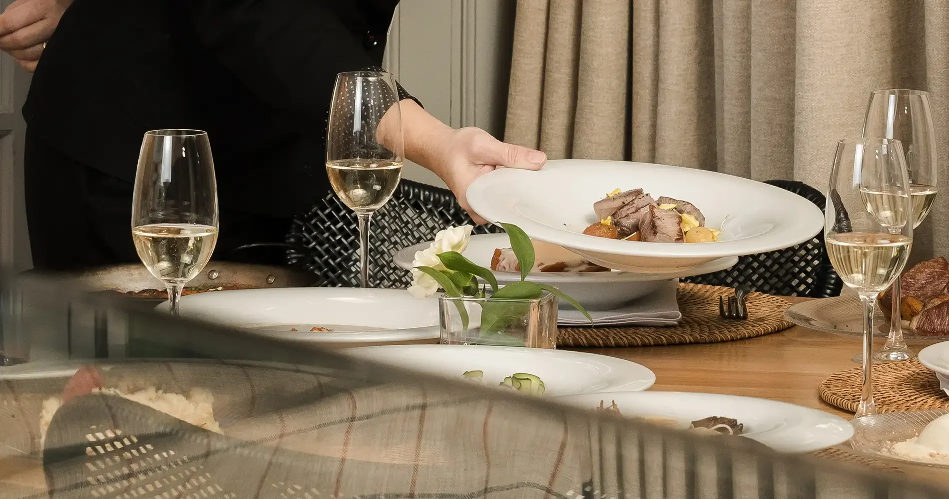 Una mano sirve comida en un plato blanco, con copas de vino en la mesa