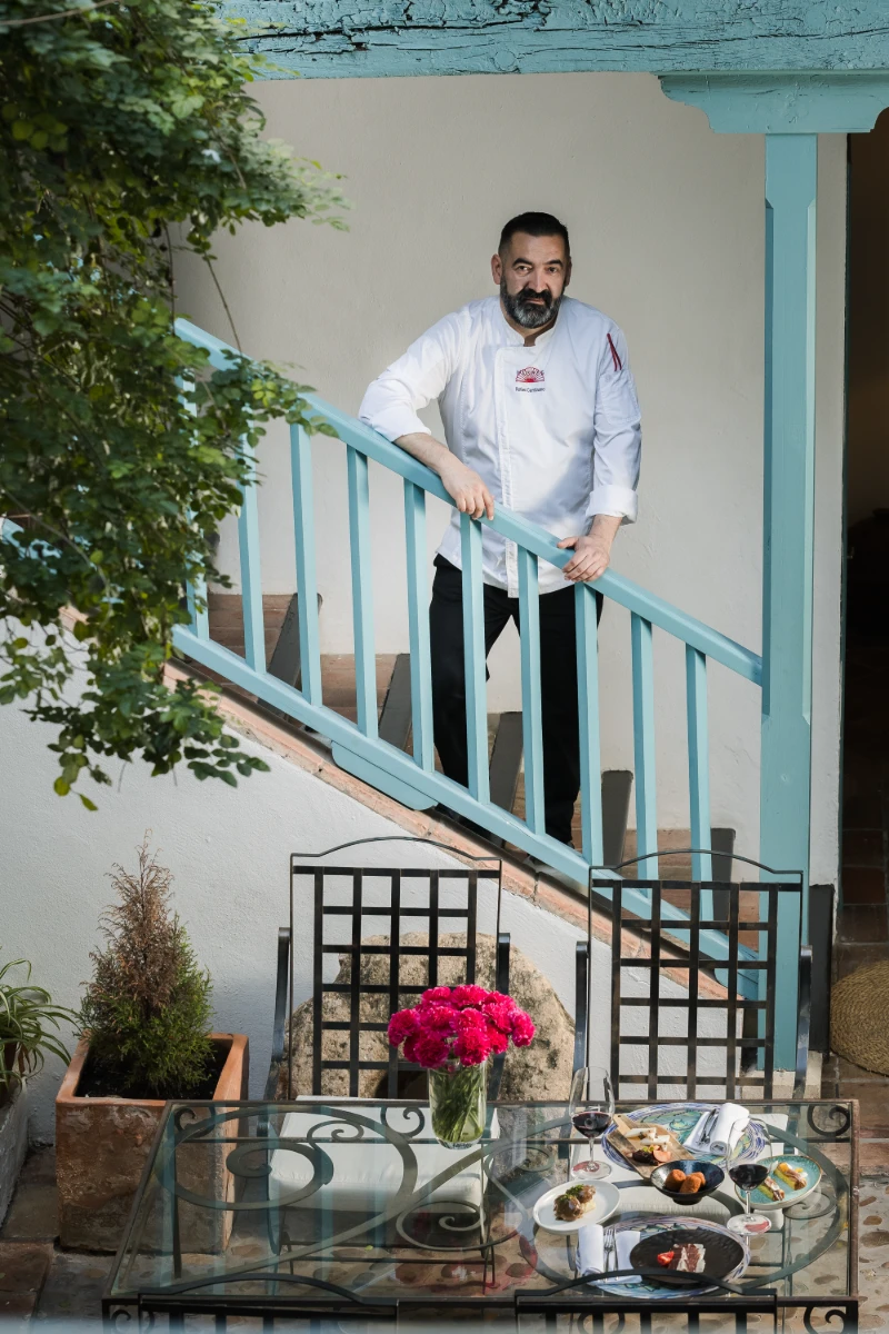Chef con barba en balc&oacute;n azul con mesa servida y flores rojas